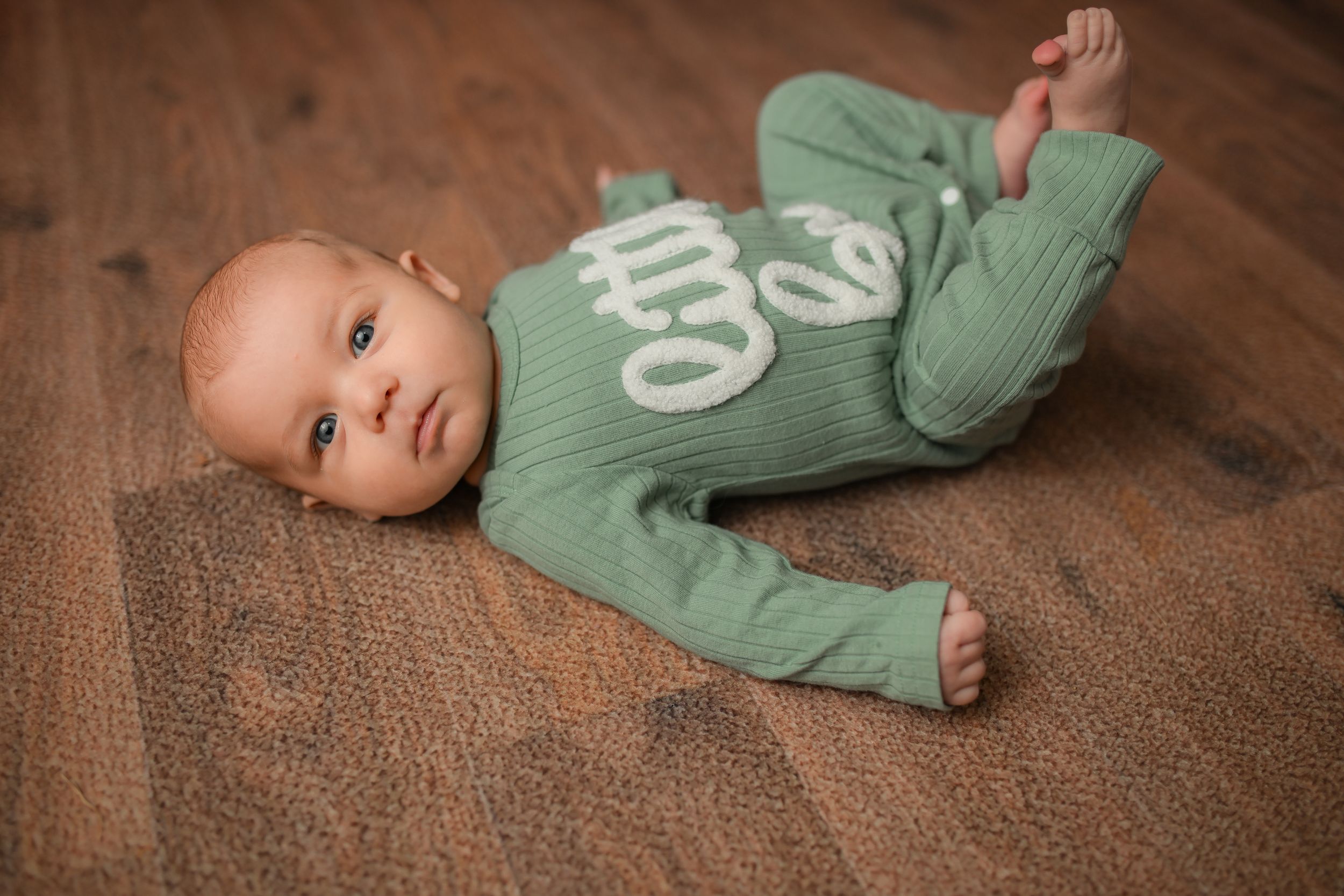 Baby in sage green onesie lying on brown textured surface during photo session.