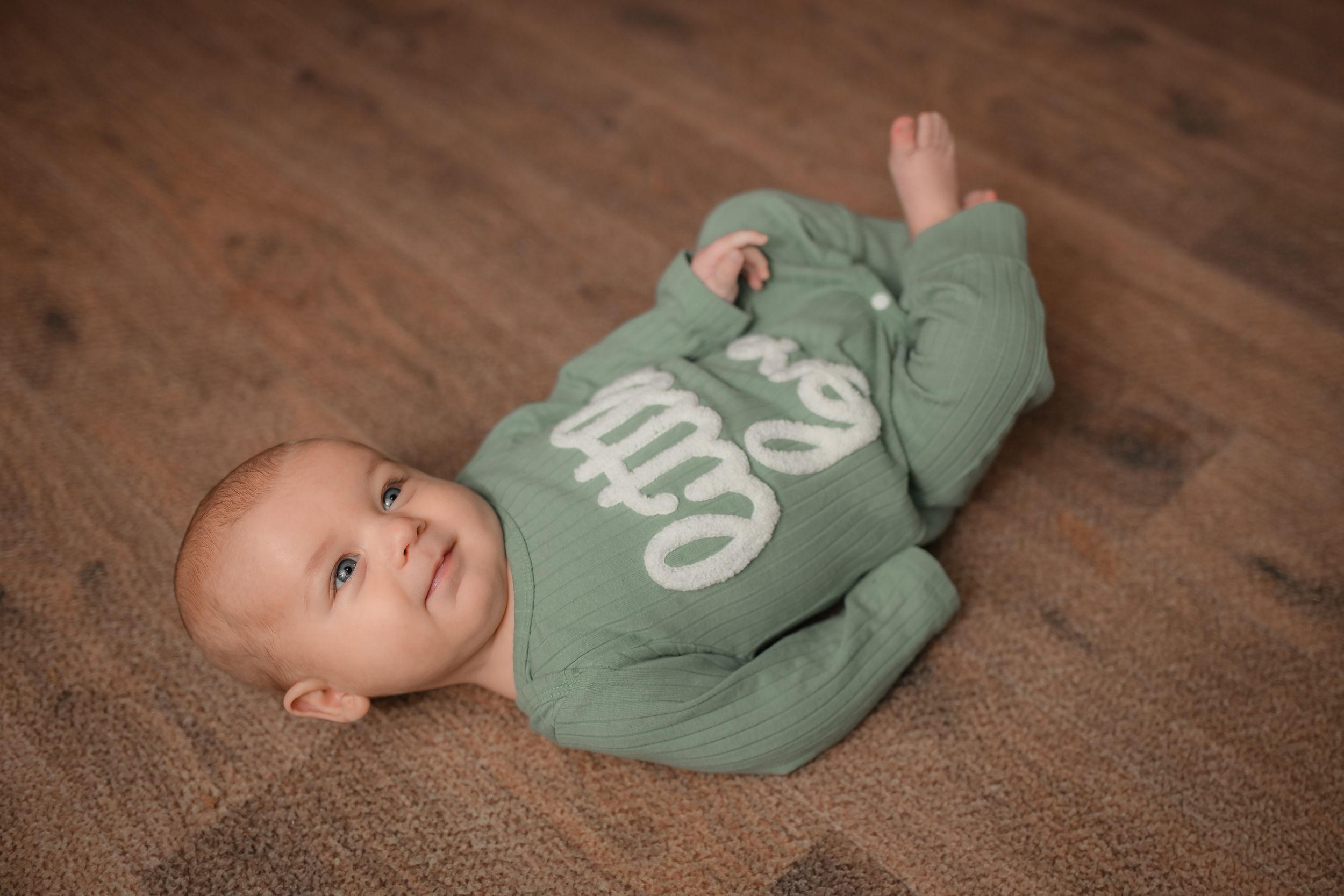 Baby in soft green sleeper lying on brown carpet during photoshoot.