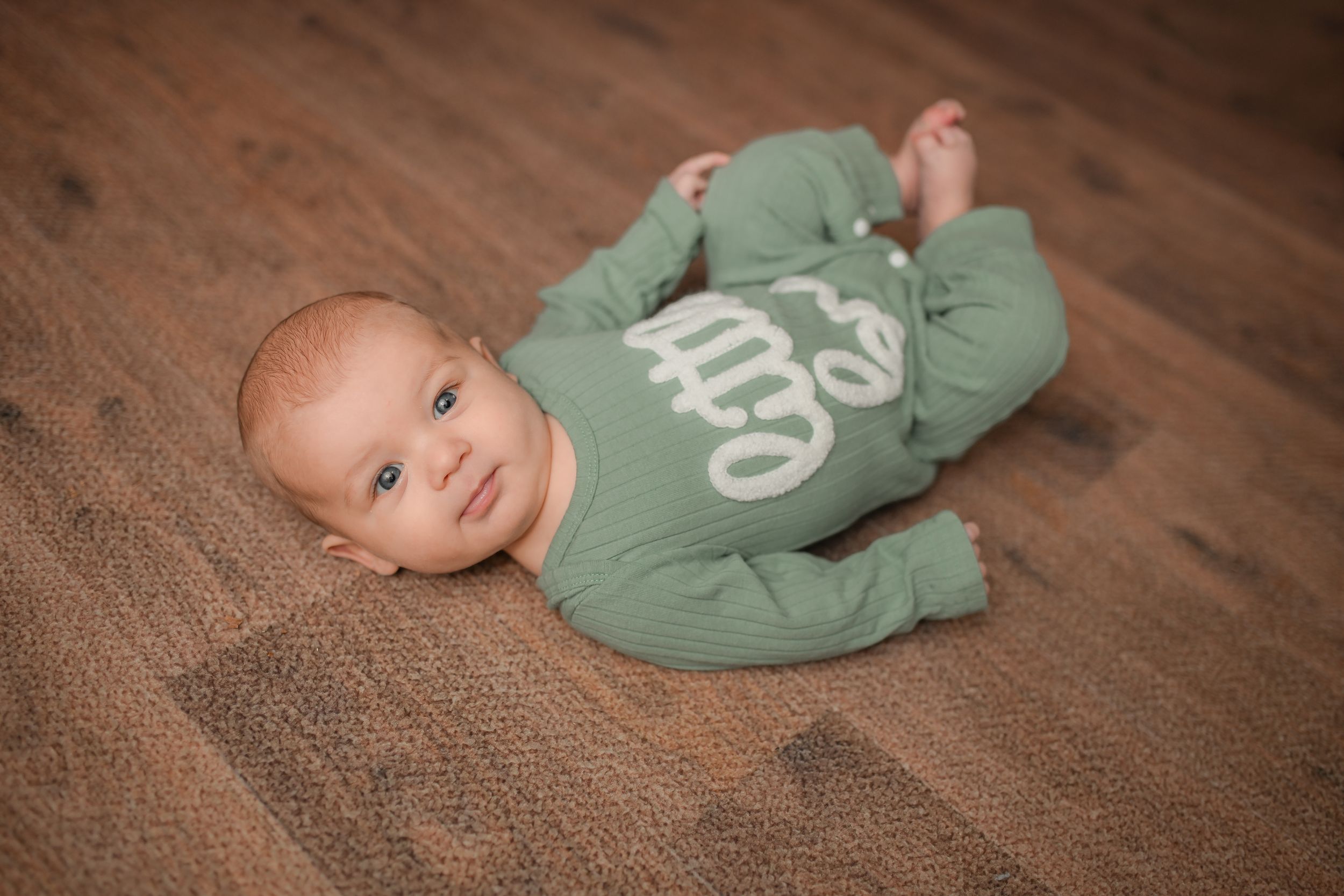 Infant wearing sage colored outfit on textured brown surface.