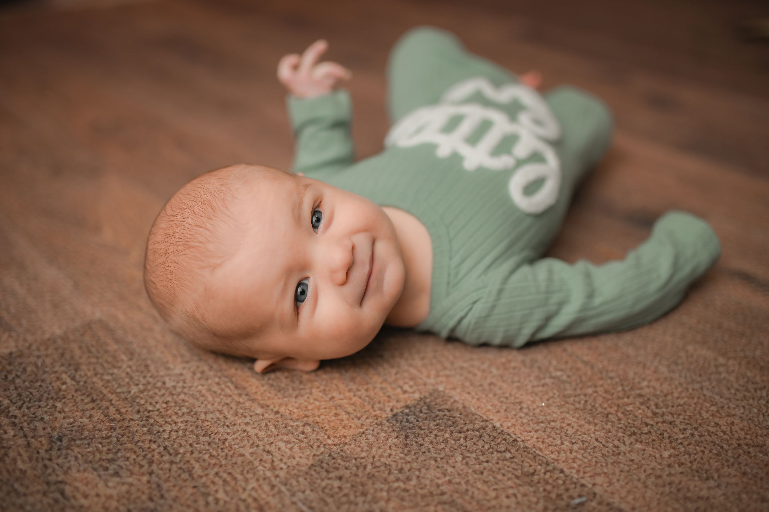 Baby in mint green sleeper lying on brown carpet with soft lighting.