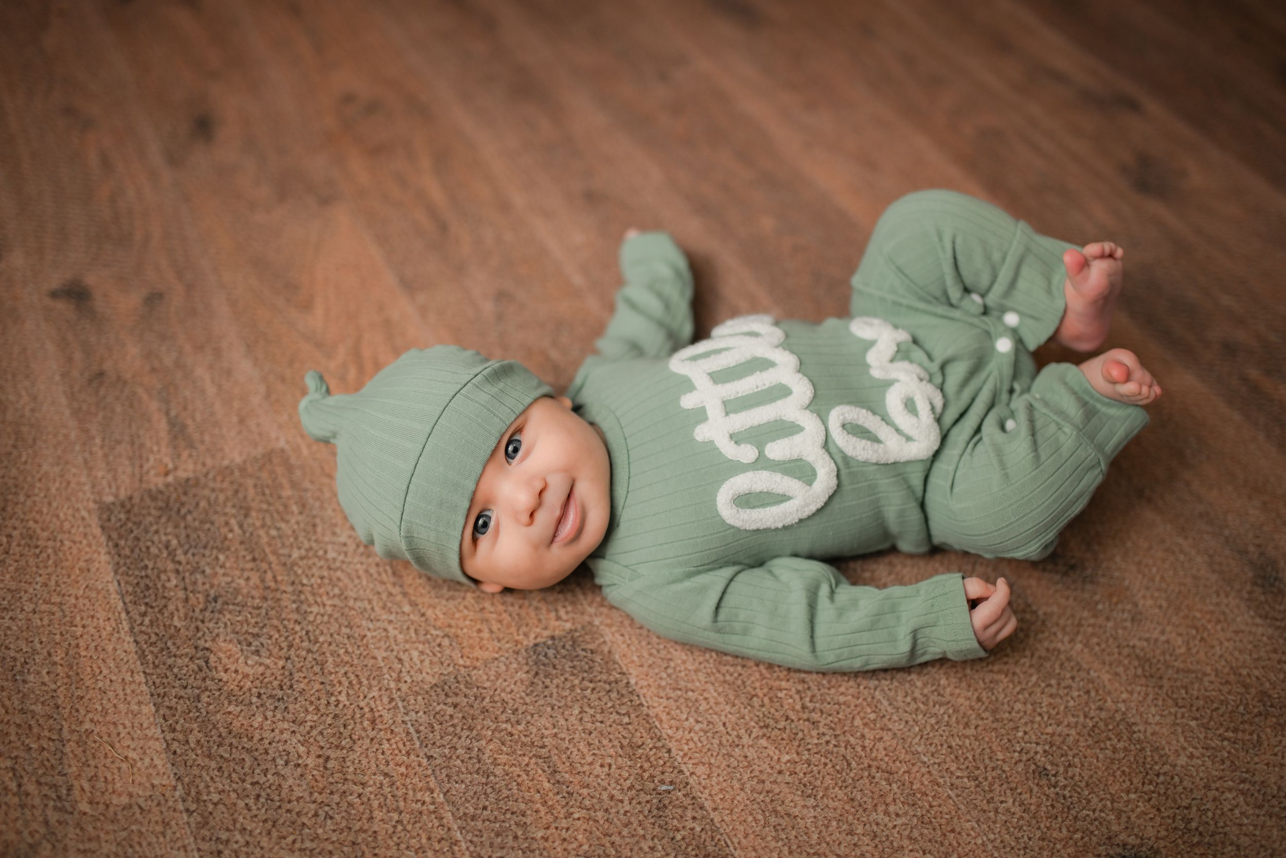 Baby in sage green outfit lying on brown textured carpet.