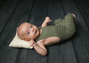 Baby in olive green outfit lying on small pillow against dark wooden floor background.