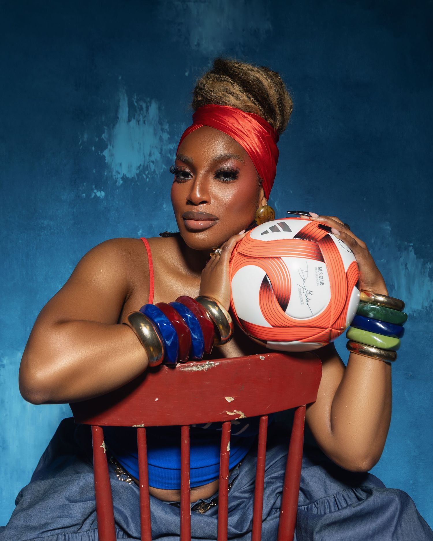 Stylish woman with colorful bangles holds Adidas soccer ball while seated on red chair.