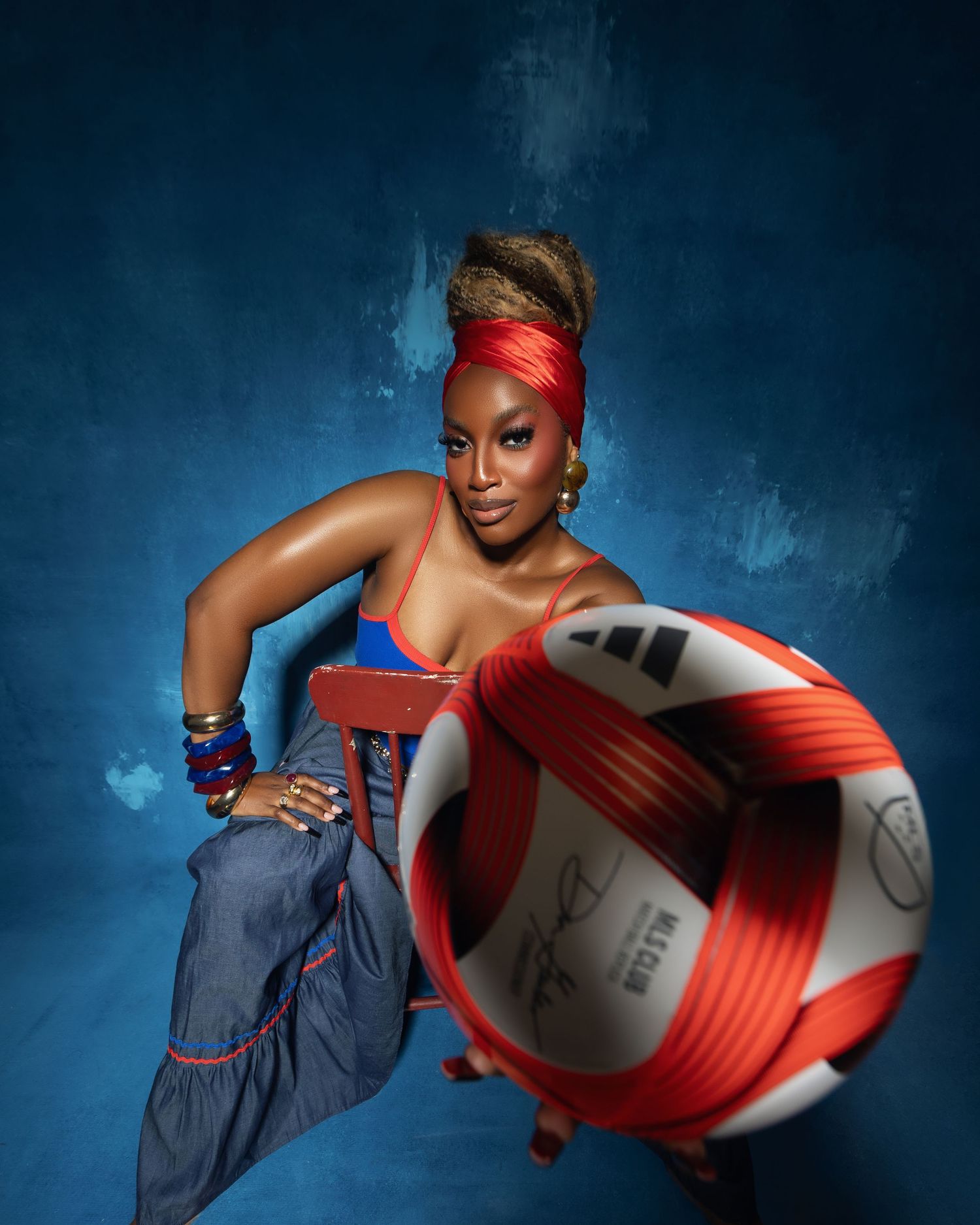 Woman in Haiti jersey and red headwrap poses dynamically with Adidas soccer ball on blue backdrop.