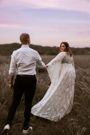 A couple in wedding attire walking hand in hand through a field at sunset, with the bride wearing a flowing lace dress.