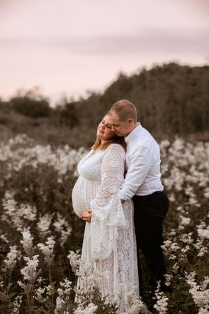 A couple shares an intimate moment in a wildflower field at sunset, with the pregnant woman wearing a white lace dress.