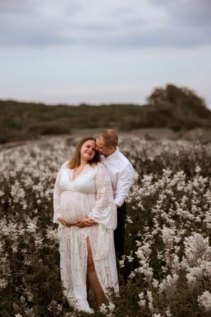 A romantic couple embraces in a field of white wildflowers during a dreamy maternity photoshoot at sunset.