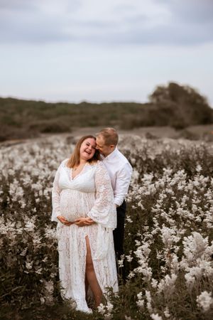 Intimate maternity portrait of a couple embracing in a field of white wildflowers under an overcast sky.
