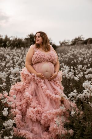 A pregnant woman wearing a flowing pink ruffled gown poses in a field of white wildflowers at dusk.