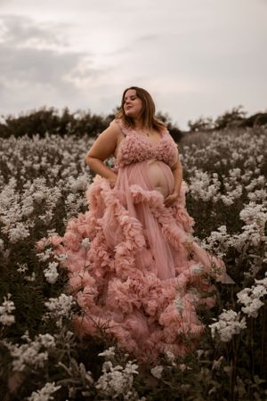 A person in a flowing pink ruffled gown poses gracefully in a field of white wildflowers at dusk.
