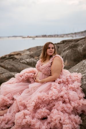 A person in a flowing pink tulle gown poses gracefully on coastal rocks against an ocean backdrop at dusk.