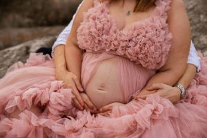 A pregnant woman wearing a flowing pink tulle maternity gown cradles her belly while sitting outdoors.