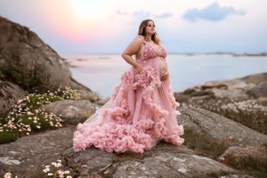 A flowing pink tulle gown with ruffles catches the sunset light on coastal rocks by the ocean.