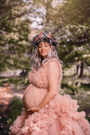 Expectant mother in pink ruffled gown and floral crown poses serenely in a dreamy outdoor woodland setting.