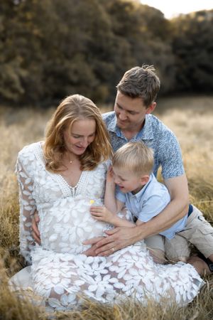 Expecting family sits together in a sunlit field while wearing white and blue outfits during a maternity photoshoot.