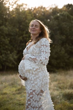 A pregnant woman in a white floral lace dress poses in a sunlit field during golden hour.