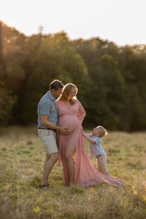 Family portrait in a golden sunset field with maternity dress and toddler touching pregnant belly.