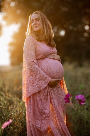 Figure in pink lace maternity dress poses in golden sunset light among wildflowers in a dreamy outdoor setting.