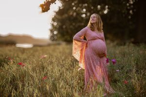 A pregnant figure in a flowing pink dress stands in a golden field at sunset with pink wildflowers blooming nearby.