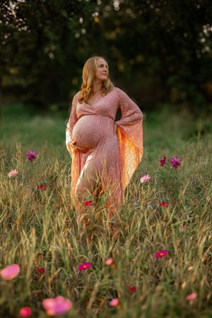 Expectant mother in flowing pink dress stands elegantly among wildflowers during golden hour sunset.
