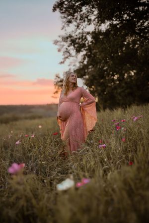 A romantic maternity photo shows a flowing pink dress in a wildflower meadow during golden hour sunset.