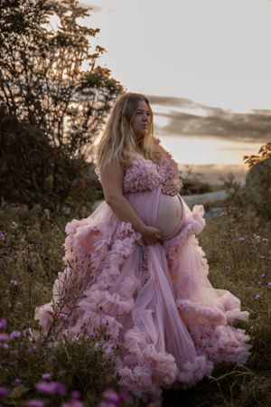 A dreamy maternity photoshoot at sunset shows an ethereal pink ruffled gown among wildflowers in nature.