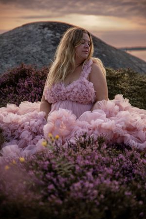 A woman in a flowing pink dress poses among purple heather flowers at sunset with mountains in the background.