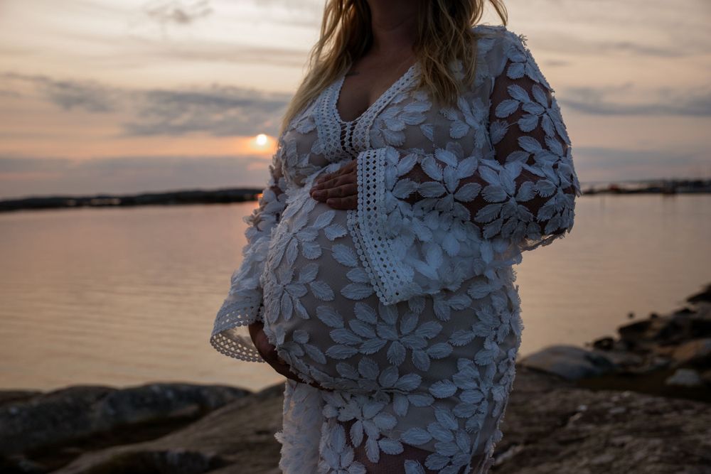 Silhouetted pregnant figure wearing a textured white lace dress stands by calm waterfront during golden sunset.