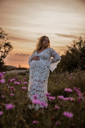 A white lace maternity dress glows at sunset in a field of pink wildflowers during a romantic outdoor photoshoot.