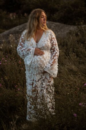 A woman in a white lace maternity dress stands in a wildflower meadow at sunset.