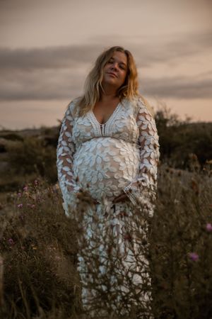 Expectant woman in white lace dress poses in field at sunset with glowing sky and natural scenery.