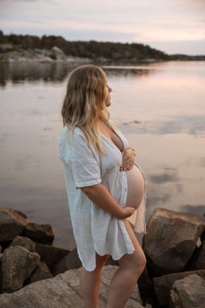 Expectant mother in white dress standing on rocky shoreline during sunset at peaceful lake.