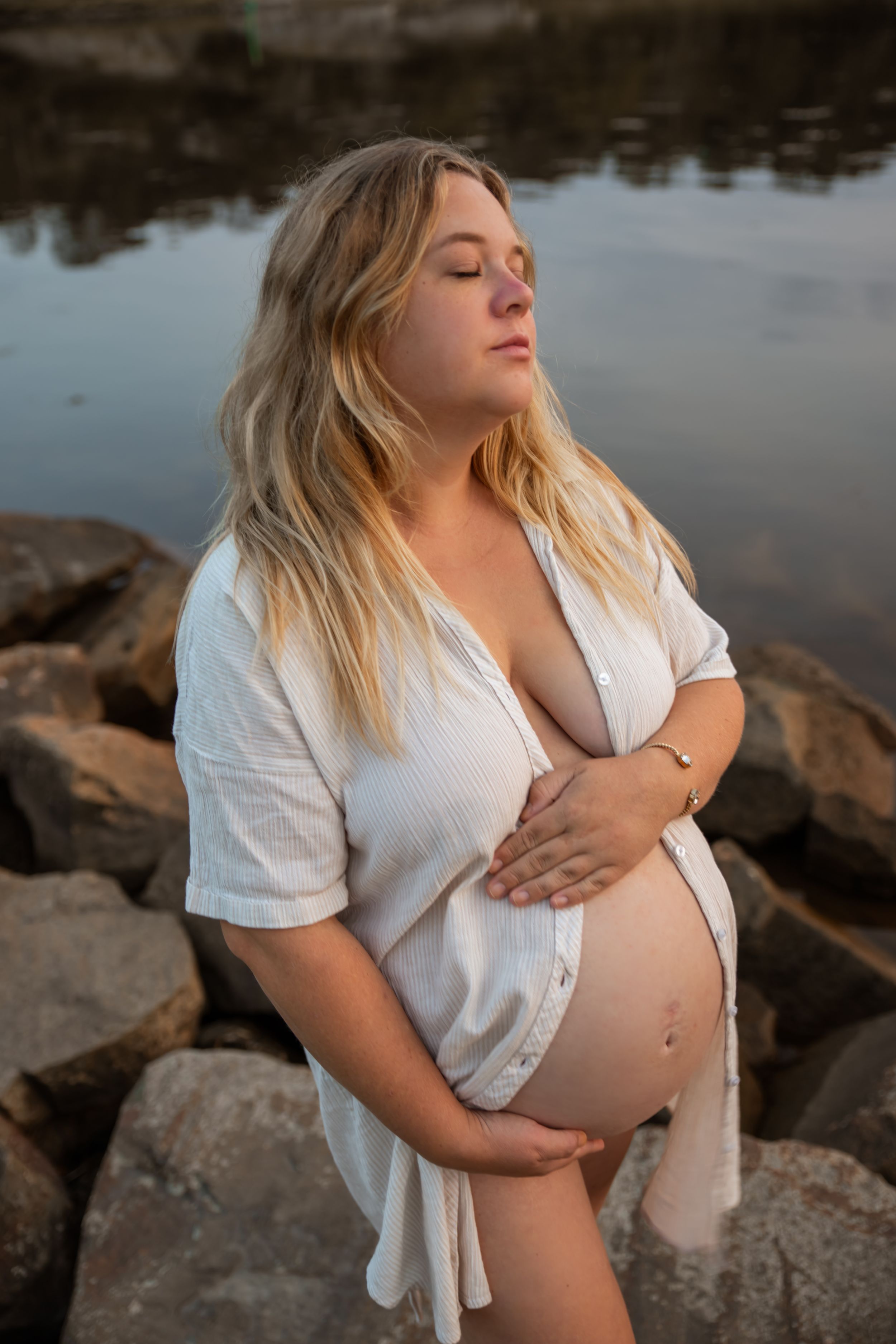A pregnant woman in a white dress poses near water during a maternity photoshoot at sunset.