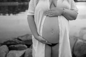 Black and white maternity photo showing pregnant belly at waters edge with rocks in foreground.