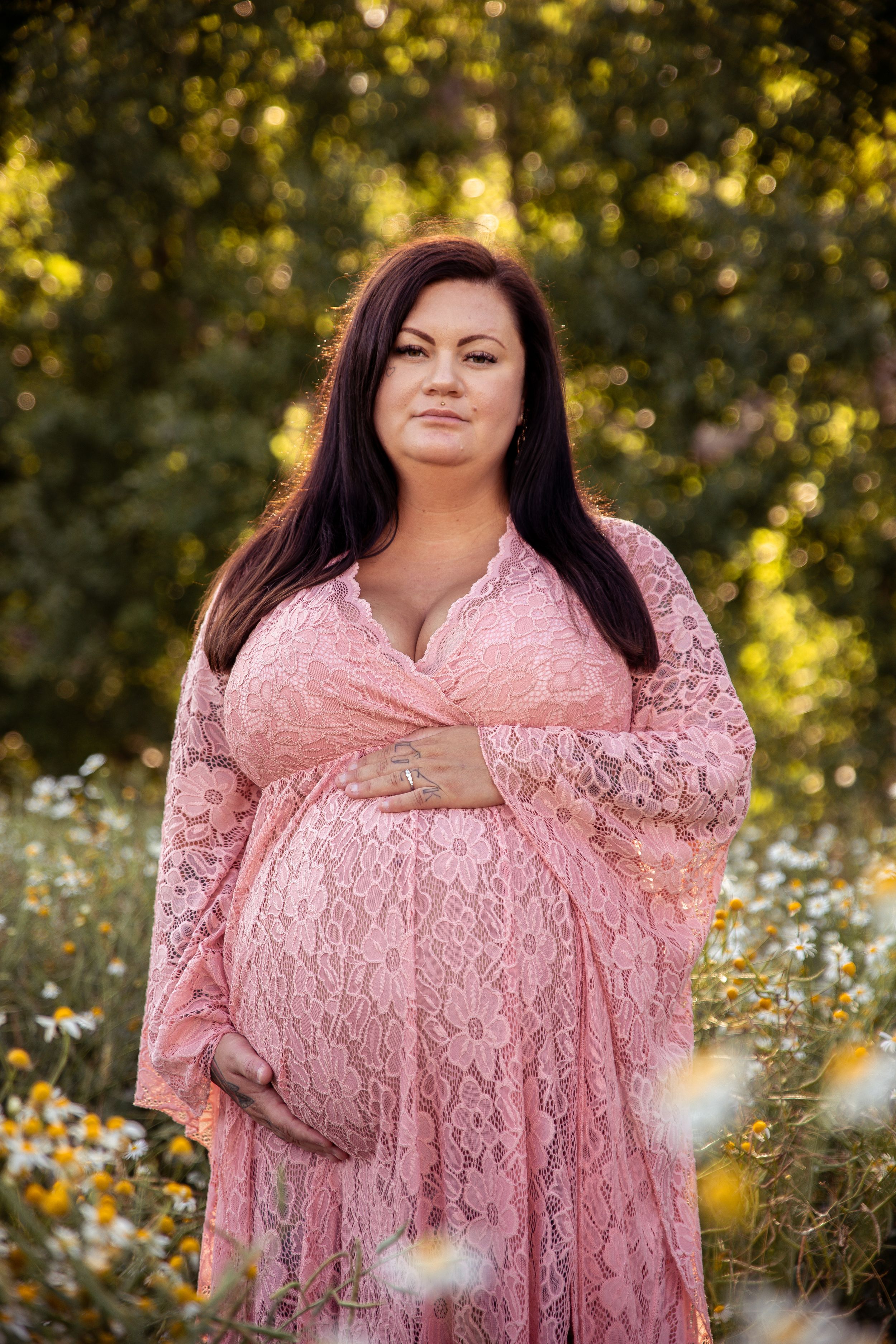 A maternity photo in a pink lace dress standing in a field of wildflowers with golden bokeh lighting.