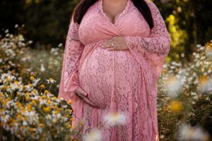 A pregnant figure in a pink lace maternity dress stands in a field of wildflowers during golden hour.