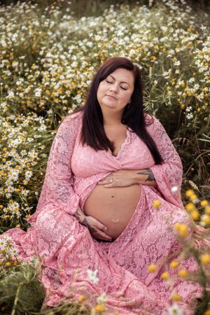 A pink lace maternity dress flows gracefully among blooming white daisies in a sunlit meadow.