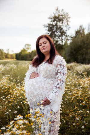 A pregnant person in a white lace dress stands in a field of blooming daisies at sunset.