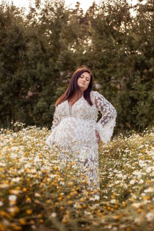 A maternity portrait in a white lace dress surrounded by blooming daisies in a garden setting.