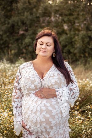 A white lace maternity dress with floral details is showcased in a golden-hour outdoor portrait session among wildflowers.