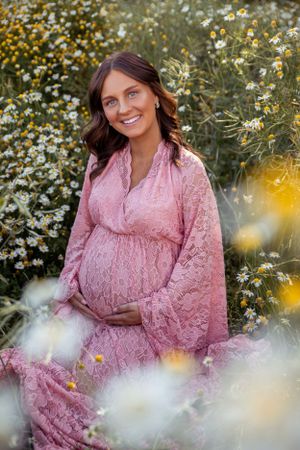 A maternity portrait featuring a pink lace dress surrounded by delicate white and yellow wildflowers in a garden setting.