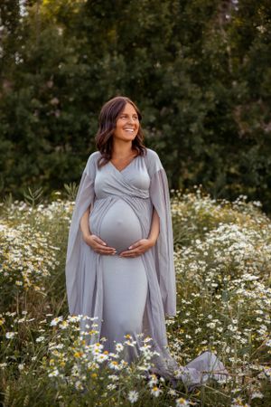 A pregnant woman in a flowing gray dress stands in a field of white daisies at sunset.