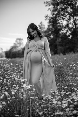 A pregnant woman in a flowing dress stands in a wildflower meadow in black and white photography.
