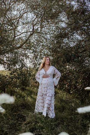 A flowing white lace dress with bell sleeves photographed in a dreamy natural setting with trees and soft lighting.