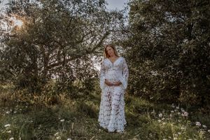 A person in a white bohemian dress stands in a sunny garden surrounded by olive trees at sunset.