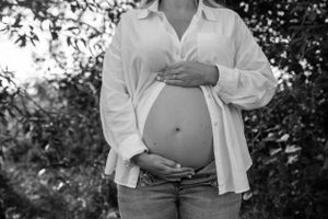 A pregnant belly is revealed beneath a white button-down shirt, against a blurred outdoor background in black and white.