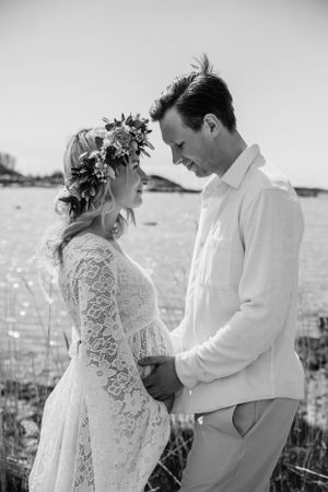 Black and white maternity photo featuring a couple in a field wearing white lace dress and casual attire with a floral crown.
