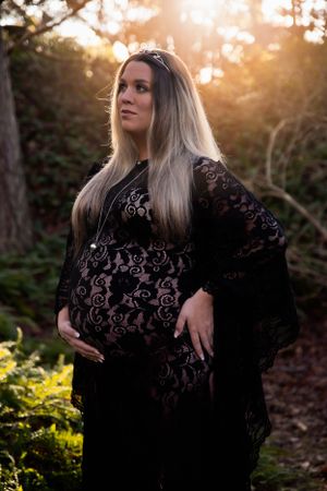 Beautiful maternity photoshoot in black lace dress at sunset with warm golden light filtering through trees.