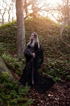 A figure in a flowing black lace dress poses dramatically in a misty forest setting at golden hour.