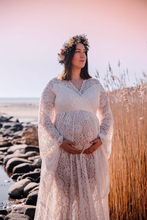 A pregnant woman in a white lace dress stands near tall beach grass at sunset wearing a floral crown.